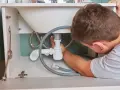 Hydraulik naprawia syfon pod zlewem, widoczne rury i węże.