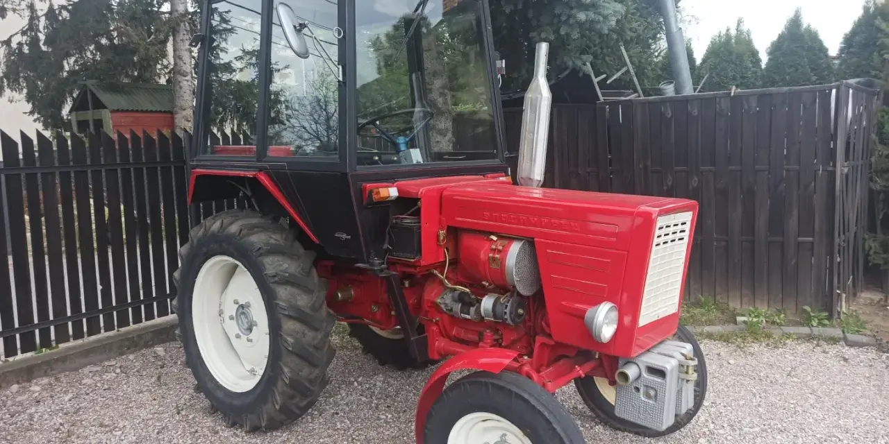 Czerwony traktor T-25 z kabiną stoi na żwirowym podwórku.