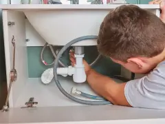 Hydraulik naprawia syfon pod zlewem, widoczne rury i węże.