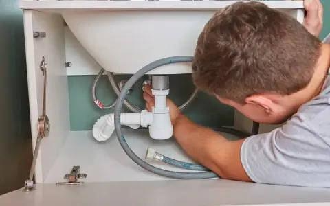 Hydraulik naprawia syfon pod zlewem, widoczne rury i węże.