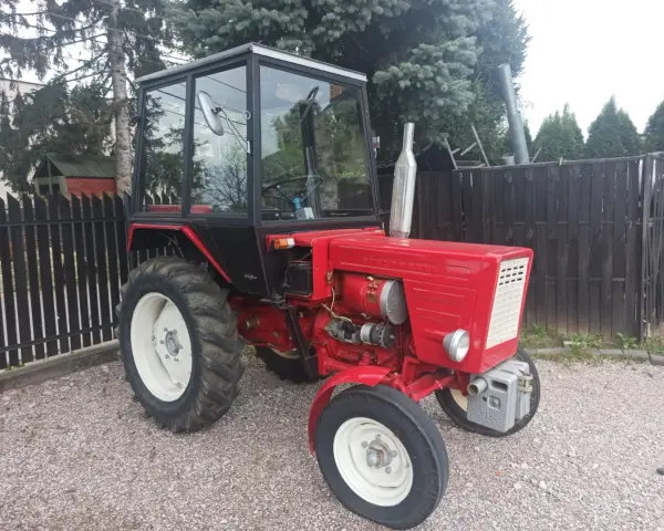 Czerwony traktor T-25 z kabiną stoi na żwirowym podwórku.