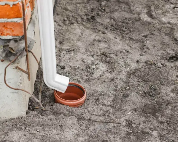 Biała rura spustowa odprowadza wodę do brązowej studzienki na ziemi.