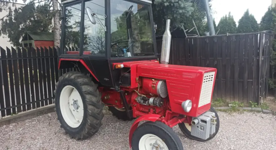 Czerwony traktor T-25 z kabiną stoi na żwirowym podwórku.