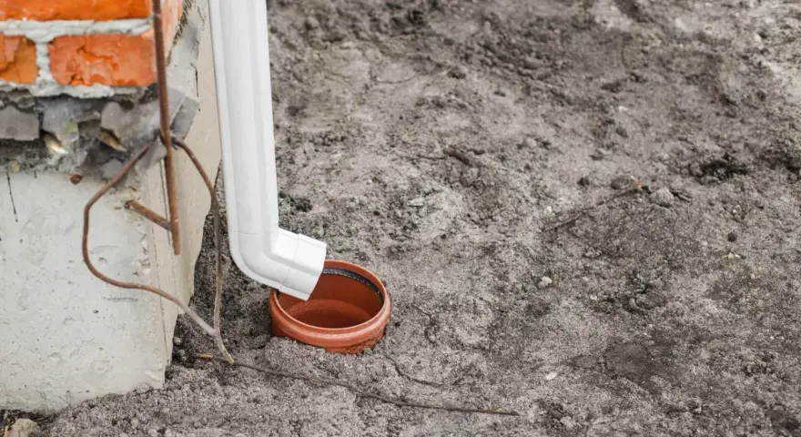 Biała rura spustowa odprowadza wodę do brązowej studzienki na ziemi.