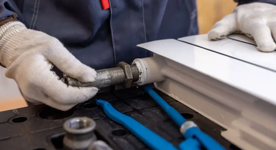Hydraulik w białych rękawiczkach dokręca metalową rurę do białego grzejnika.