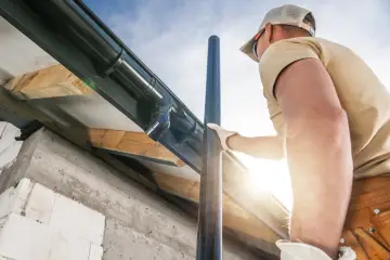 Mężczyzna w rękawiczkach montuje rynnę i rurę spustową na tle nieba.